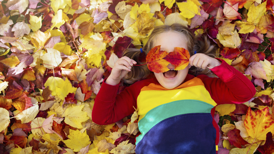 En flicka ligger på en bädd av färgglada höstlöv. Hon skrattar och håller upp två löv för ögonen.