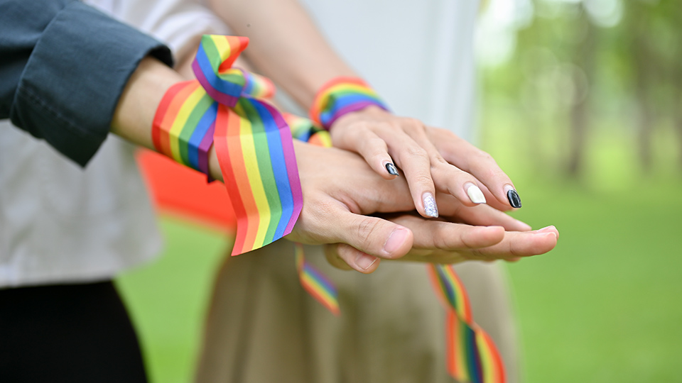 Tre personer med regnbågsfärgade band runt handlederna håller ihop sina händer.