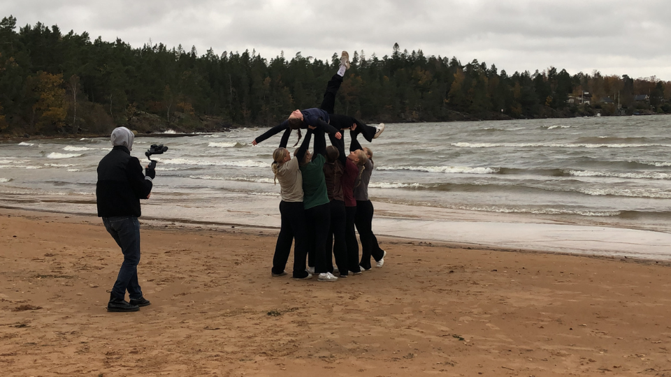 Bilden är vid en strand. På stranden står ett gäng ungdomar tätt till varandra och lyfter upp en annan tjej i luften. Bredvid gruppen är en filmskapare som filmar med kamera. 