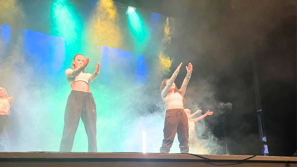 En bild med fyra dansare på en scen. Två dansare står baktill på scenen och två dansare framtill. Samtliga sträcker ut sina armar. I bakgrunden ser man rök som lyser i olika färger. 