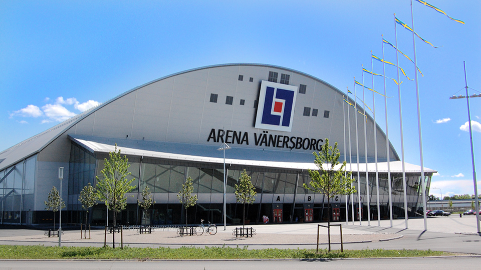 Arena Vänersborg från håll mot en blå himmel och sverigevimplar på flaggstängerna. 