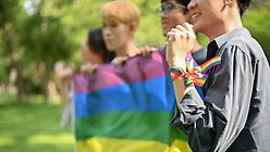 Fyra unga personer håller i en regnbågsflagga. Personen närmast kameran har ett regnbågsband runt handleden.