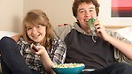 En ung kille och tjej sitter och tittar på TV med en skål popcorn framför sig.